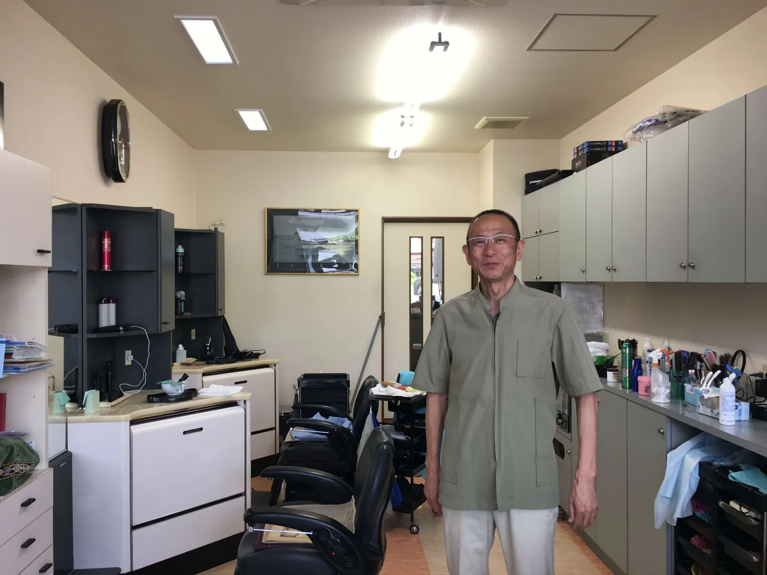 明るく近代的な店内で、理容椅子と戸棚のあいだに理容師が立っている。