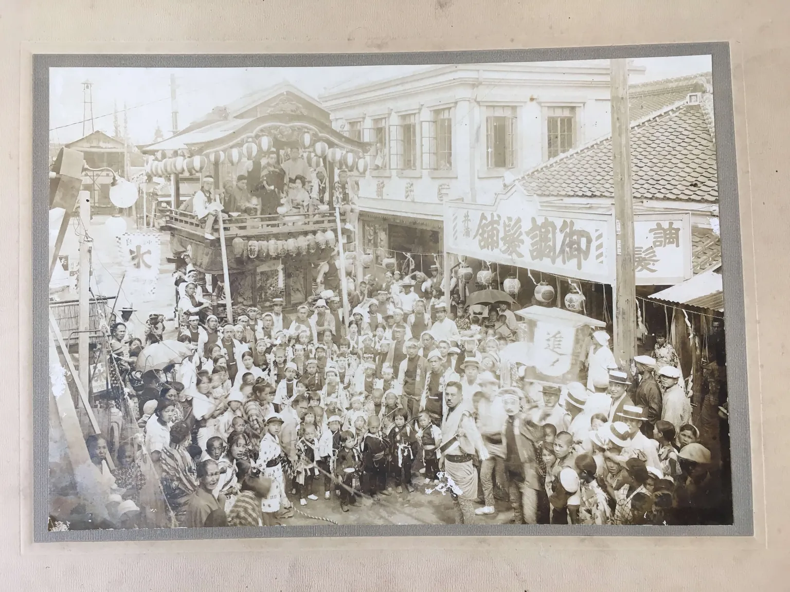 古いセピア色の写真。旗の下がる二階建ての店が並ぶ通りに、祭りの人だかりができている。