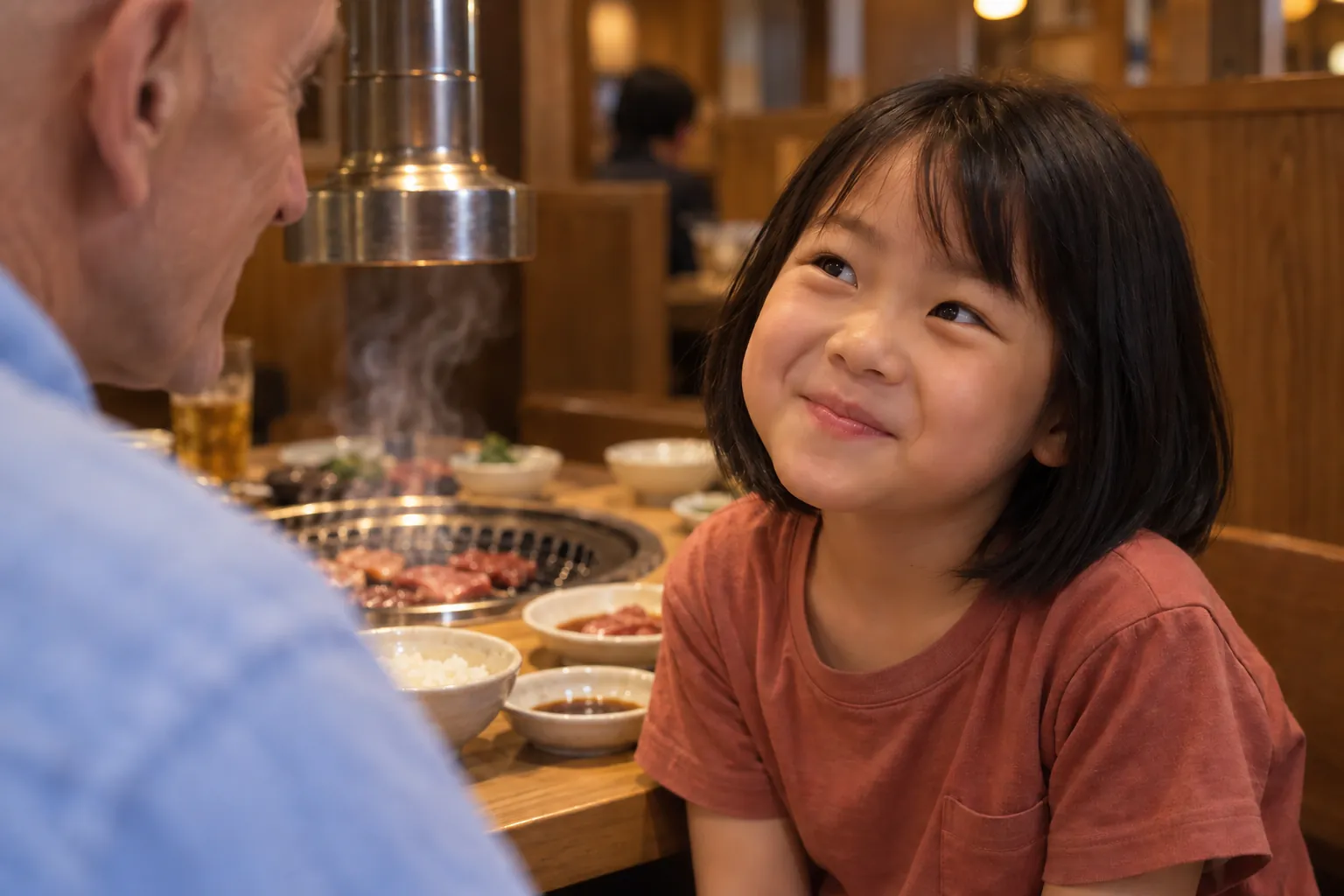 A bob-haired Japanese girl smiles up at a bald man in a blue shirt, with grilled meat and rising steam behind her.