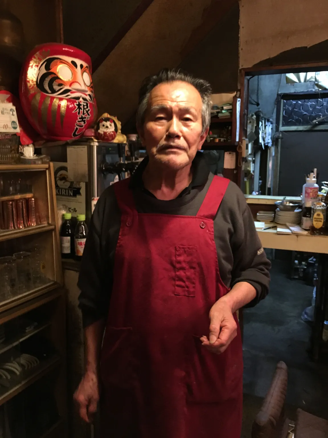 赤いだるまの下、赤い前掛け姿で調理場に立つ「ねぎし」のマスター。