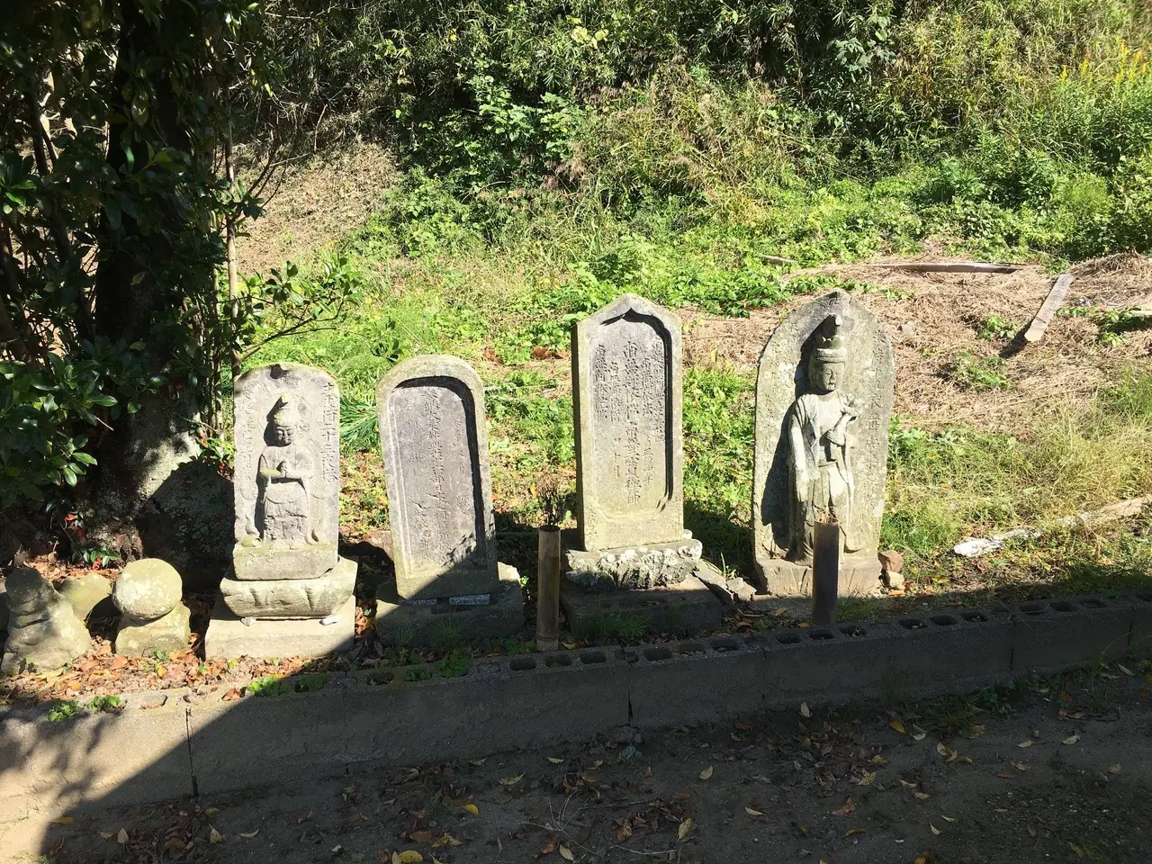 日の当たる草の土手に並ぶ、風化した石の神さまと石碑。